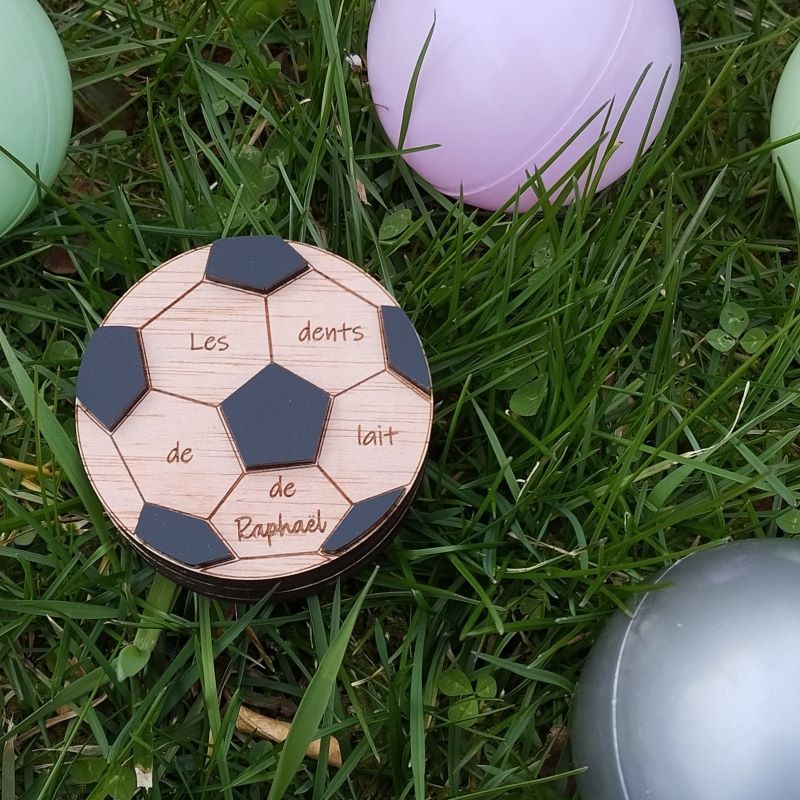 boite a dents en forme de ballon de football. le prénom de l'entant est gravé desus après la mention "les dents de lait de...." Le couvercle est orné de plexiglas noir mat.